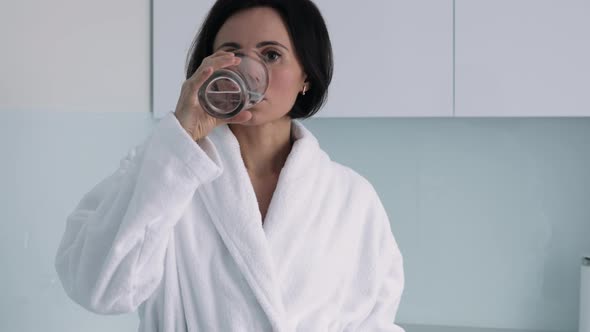 Thirsty smiling 35s woman in white bathrobe drinking water