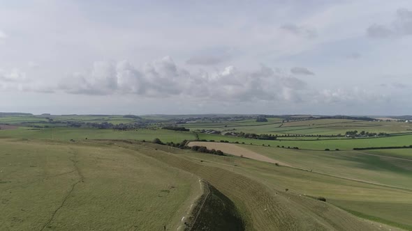 Aerial tracking slowly west above the northern ramparts of Maiden Castle. Beautiful green fields and