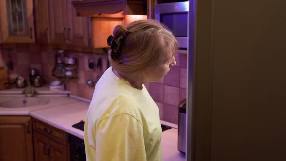 Eating disorder, hungry middle aged woman opens a refrigerator at night and eating unhealthy food.