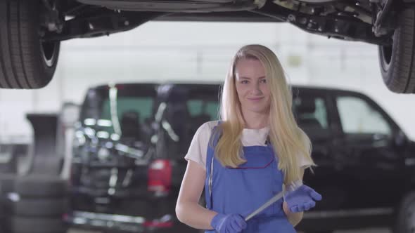 Portrait of Young Female Auto Mechanic in Workrobe Standing Under Car in Repair Shop