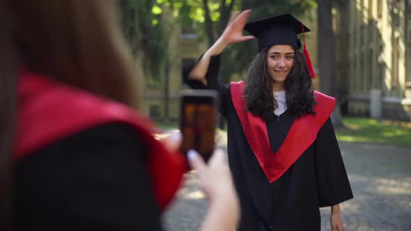 Cheerful Graduate Woman Throwing Mortarboard Cap As Friend Taking Photo on Smartphone