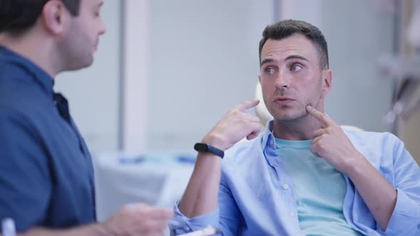 Portrait of Handsome Caucasian Man Talking with Middle Eastern Doctor Sitting in Dentist Chair in