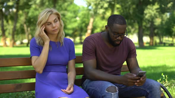 African American Man Playing Cellphone on Date in Park, Ignoring Sad Girlfriend