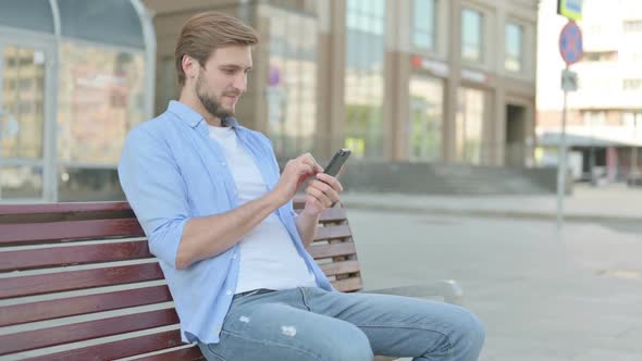 Man Celebrating Online Success on Smartphone While Sitting Outdoor on Bench