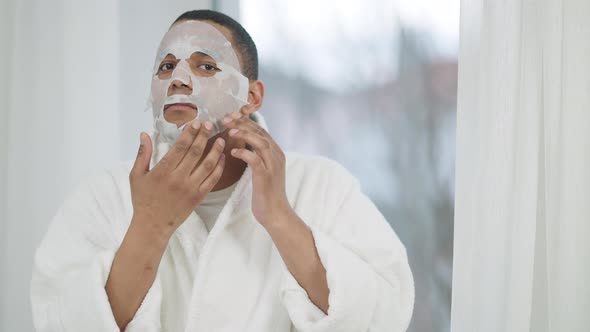 Mirror POV of Young African American Man in White Bathrobe Putting on Purifying Facial Mask Standing