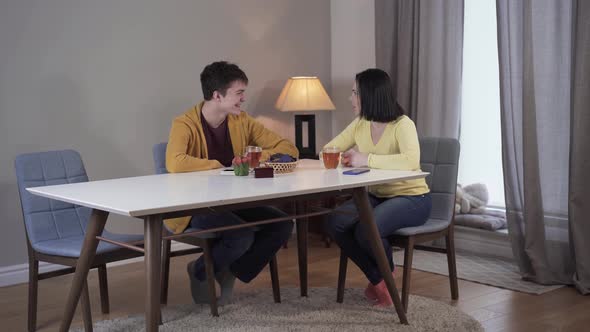 Long Shot of Happy Caucasian Teenage Son and Adult Mother Sitting at the Table and Talking, Smiling