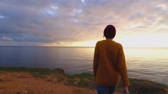 Young GoodLooking Woman Wearing Red Hat and Yellow Sweater and Slowly Approaching Ocean