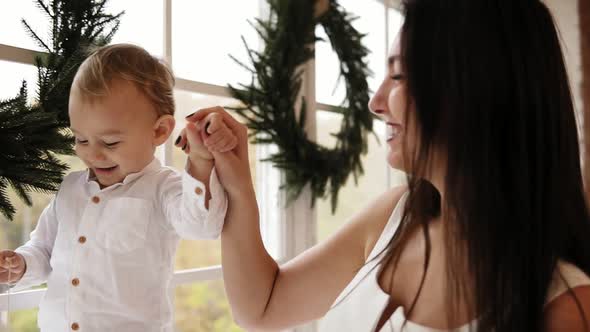 Young Mother is Holding Her Son's Hands While He is Walking on the Window Sill Decorated with