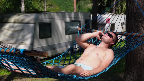 The Man is Resting Lying on a Hammock in the Woods He is Rocking and Talking on a Smartphone