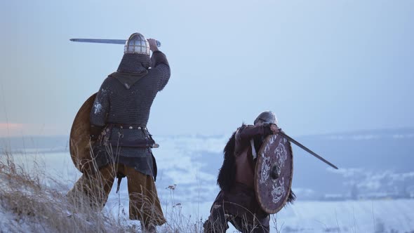 Two viking are fighting with axes and shields on the winter meadow.