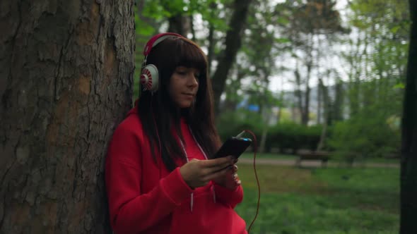 Woman, standing near a tree in the park, uses a smartphone and listens to music with headphones.