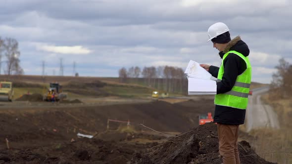 The Worker Looks at the Drawings on the Background of the Road Under Construction