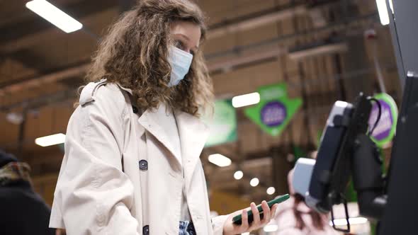Woman in a Medical Mask Makes Purchases at the Selfservice Checkout in a Supermarket Using a Mobile