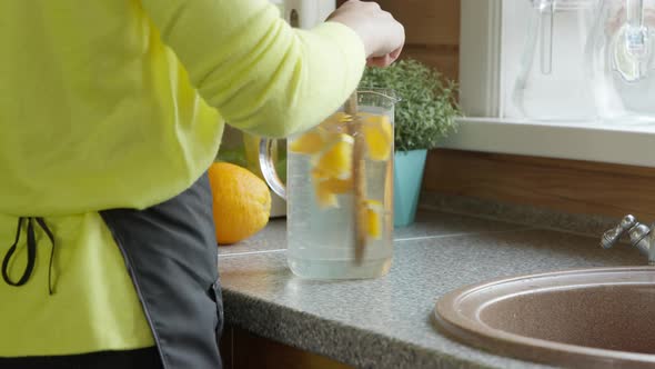 Young Caucasian Girl Stirs a Wooden Spoon with Citrus Lemonade in the Kitchen