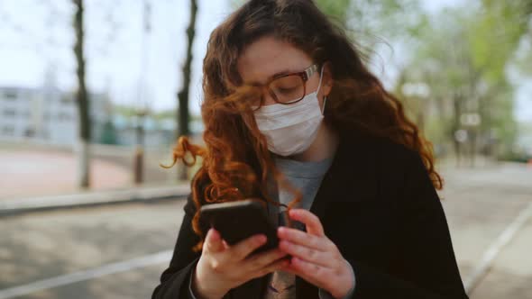 Young Girl in a Medical Mask Uses a Smartphone in the Park