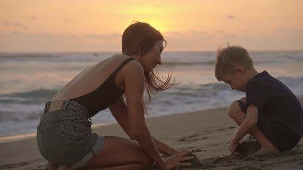 Mother and Little Son Playing with Sand on Beach at Sunset