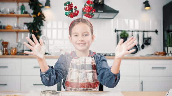 Cute Little Girl Wearing Apron and Funny Deer Horns Clapping Hands with Flour Laughing to Camera at