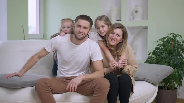 Happy Caucasian Father Mother and Daughters Looking at Camera Smiling Sitting on Couch in Living