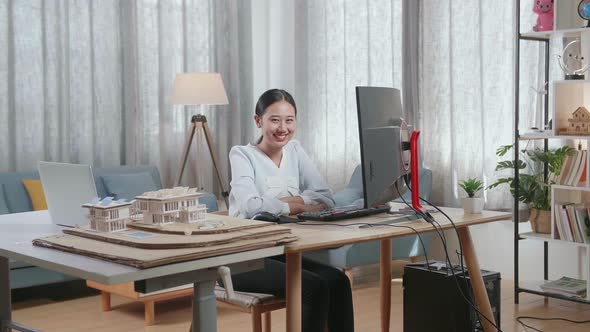 Asian Woman Engineer With The House Model Smiling To The Camera While Working On A Desktop At Home