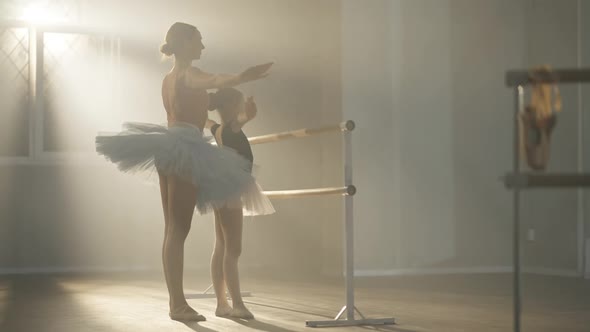 Caucasian Ballet Teacher Explaining Hands Movement to Talented Girl in Dancing Studio in Backlit Fog
