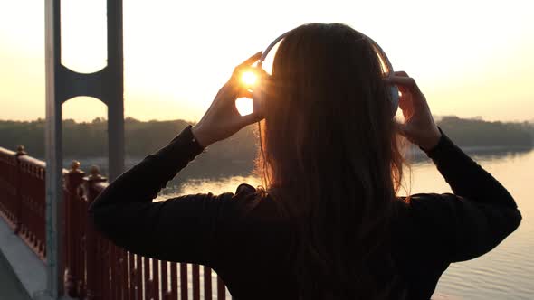 Silhouette of Pretty Woman Putting on the Headphones, Listening To Music on the Bridge. Sunset Light