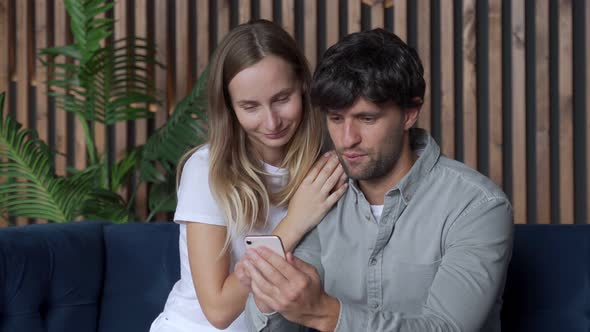 Smiling Young Couple Using Smartphone Together on Couch at Home