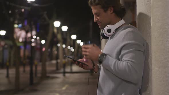 Caucasian male wearing headphones, drinking, using his phone in the evening