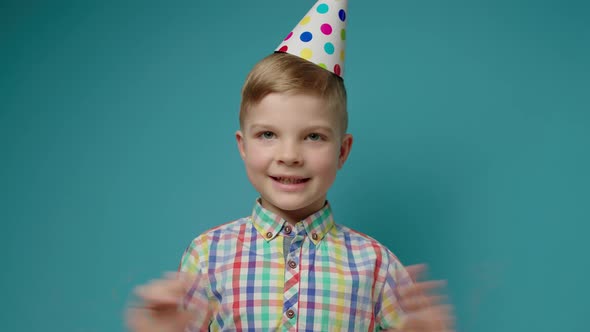 Smiling Preschool Boy in Birthday Party Hat Waving Hands and Talking to Camera on Blue Background