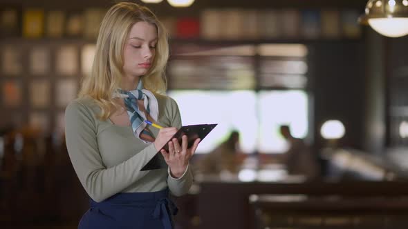 Concentrated Young Woman Writing Order with Pen Serving Customers in Restaurant Indoors