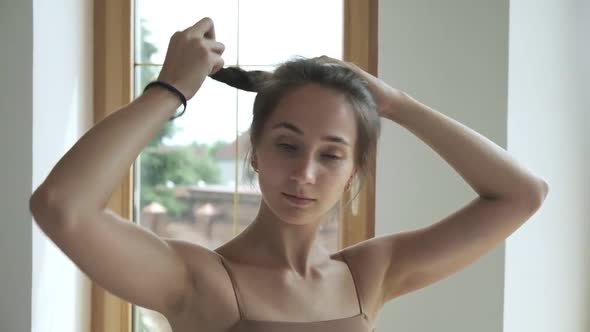 Sporty Brunette Pretty Female Stands at Window Fixes Hair Collects Hair in Bun