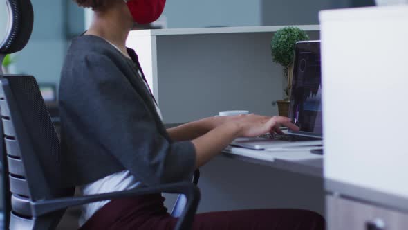 Mixed race woman wearing face mask in office, using laptop