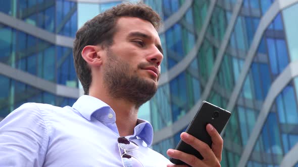 A Young Handsome Businessman Works on a Smartphone, Closeup From Below, an Office Building