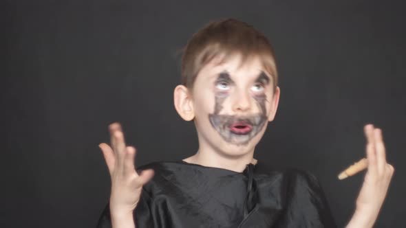 A playful caucasian boy in a zombie makeup and carnival black clothes looks at the camera with a fri