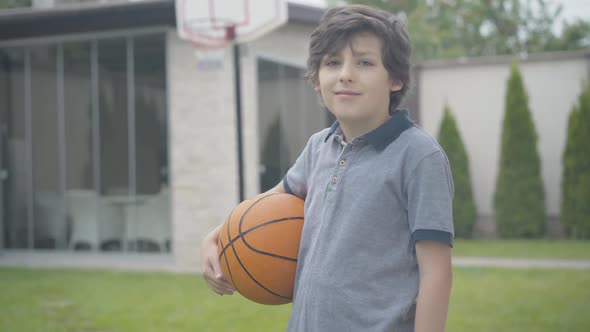 Portrait of Confident Brunette Boy Posing with Basketball Ball Outdoors. Cute Caucasian Schoolboy