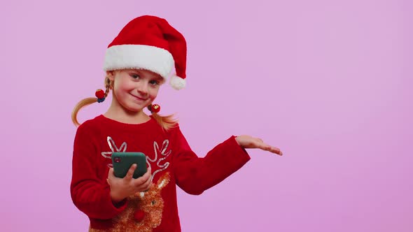 Girl Toddler in Christmas Santa Hat with Mobile Phone Showing Pointing Empty Place Advertising Area