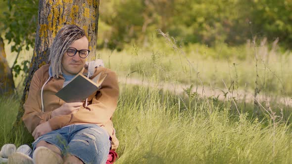 A Man in Casual Clothes and Glasses Reads Literature Under a Tree