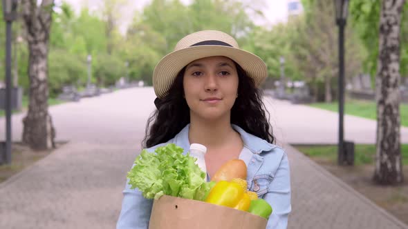 A Young Attractive Woman in a Denim Jacket and Hat Carries a Grocery Bag While Having a Good Mood