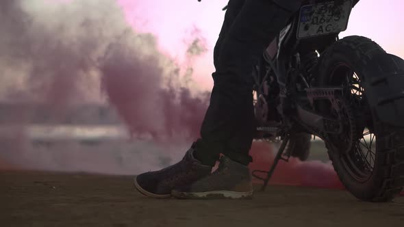 The Biker is Leaning on His Bike While the Engine is Running and Blowing Out Colored Exhaust Smoke