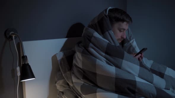 Young Man Cell Phone Addict Wakes Up Late at Night in Bed Wrapped Upside Down in Blanket Using