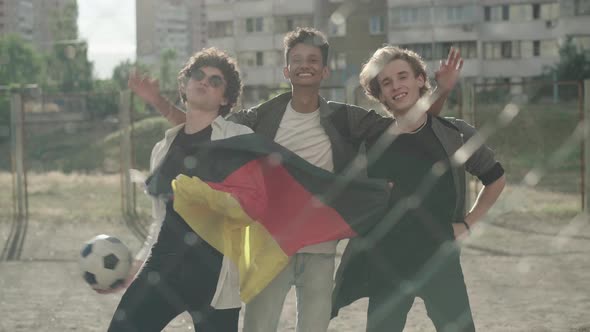 Three Happy Multiethnic Friends Posing with Sport Ball and German Flag Behind Mesh Fence. Portrait