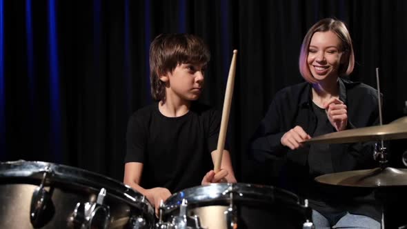 Young Caucasian Woman Teaches a Boy to Play the Drums in the Studio on a Black Background alt