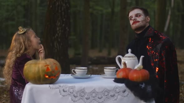Thoughtful Little Girl and Man in Vampire Costume with Face Paint Sitting at Table in Forest