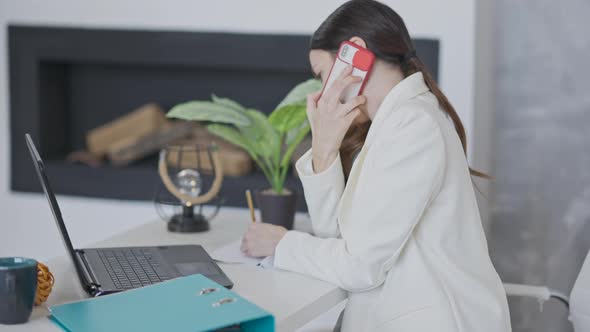 Side View of Busy Young Employee Talking on the Phone and Surfing Internet on Laptop Sitting in