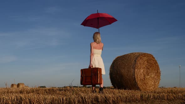 blonde girl with suitcase and umbrella is staying by a rolled haystack in field in sunset time