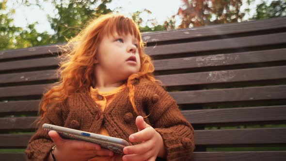 Toddler Girl with Down Syndrome Sitting on the Bench and Playing on Smartphone