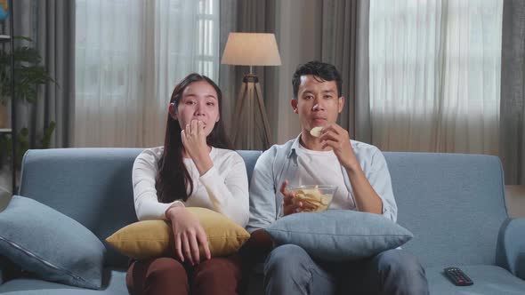 Young Asian Couple Eating Potato Chips And Watching Tv On Couch In Living Room