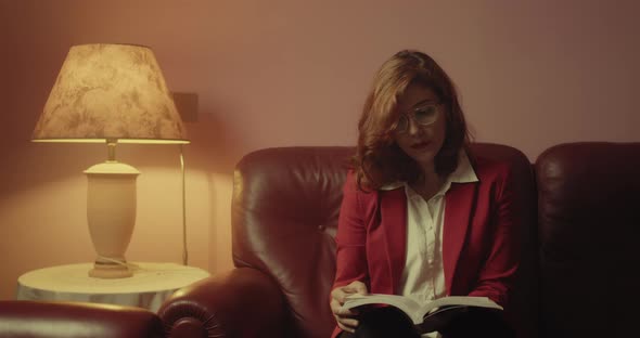 Beautiful woman with red jacket reads the book sitting on the sofa at home with eyeglasses