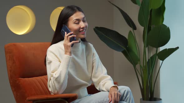 Smiling Young Woman Talks on a Smartphone While Sitting on a Chair at Home