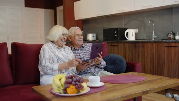 Senior Pensioner Couple with Digital Laptop Pc Computer at Home. Resting on Sofa in Cozy Living Room