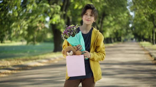 Confident Handsome Teenage Boy Standing in Summer Park with Gift Bag and Bouquet of Flowers